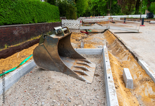 Large excavator bucket rests on gravel at a road construction site, with sand trenches, concrete curbs, temporary barriers, brick wall, and urban infrastructure work.