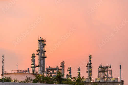 Obraz na plátne Refinery towers and tanks rise above greenery beneath a soft pink sunset, showcasing an industrial petrochemical complex with pipelines and equipment