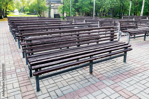 Rows of empty wooden benches with metal frames stand on a paved plaza in a green city park. Trees and grass surround the area under a cloudy sky. Public seating and outdoor gathering space concept.