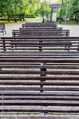 Wooden slats of park benches arranged in neat rows on a paved plaza. Green trees and a monument in the background. Public seating and outdoor recreation concept.