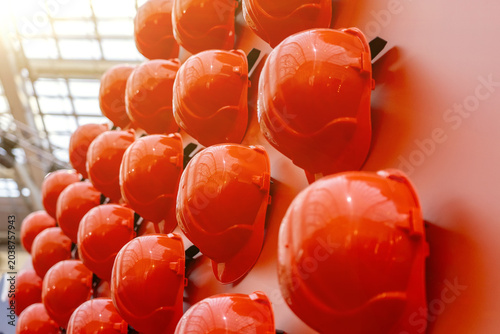 Collection of bright orange construction safety hard hats arranged in rows on wall, symbolizing industrial work, protection, safety standards, and engineering infrastructure development.