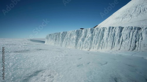 A dramatic Antarctic landscape dominated by a towering ice wall beneath a bright, clear blue sky.