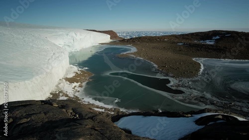 Antarctic scenery featuring an oasis with lakes and rocky terrain, with stones scattered across the landscape beneath a clear blue sky.