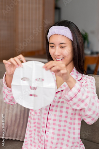 young Asian woman applying facial sheet mask smiling happy enjoying morning skincare routine beauty treatment at home bedroom wearing pink pajamas headband