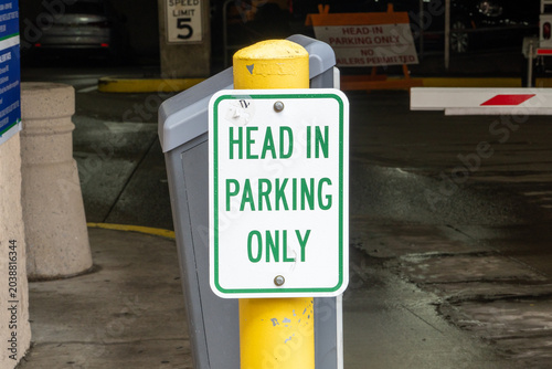 Fotografía Detailed 'Head In Parking Only' sign on a vibrant yellow pole, featuring raindrops from a recent shower, situated within a concrete parking garage environment emphasizing vehicle traffic regulations