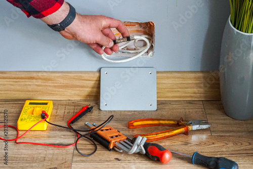 Foto A man's hand pulling a white coaxial cable from a wall mounting hole, with installation tools like a multimeter, pliers, screwdriver, and allen keys lying on the floor during electrical works