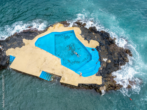Aerial view of Piscina de Caloura with a person swimming in turquoise water surrounded by volcanic rocks and crashing ocean waves in Agua de Pau, Acores, Portugal.