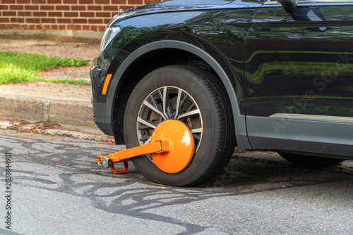 Foto Black car with bright orange wheel clamp (boot) immobilized on an urban street curb, indicating a parking violation, fine, or unpaid tickets