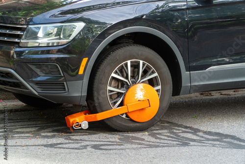 Tableau sur toile Black SUV with bright orange wheel clamp on front tire, signifying parking violation, fine, or impoundment in an urban street