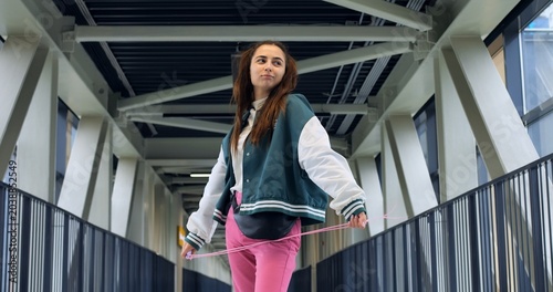 Fotografía Caucasian woman standing center on bridge wearing teal varsity jacket and pink t