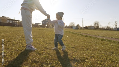 Mother and daughter holding hands, walking through sunlit green field on a warm spring day, sharing playful bonding, joy and quiet connection in the countryside
