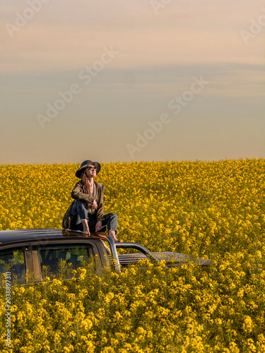 Thoughtful Woman Sitting on Pickup Truck Roof in Blooming Rapeseed Field