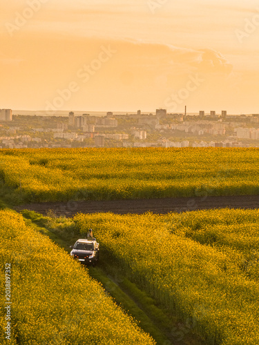 Woman Sitting on Pickup Truck Roof in Rapeseed Field at Sunset With City Skyline Background Vertical Aerial View