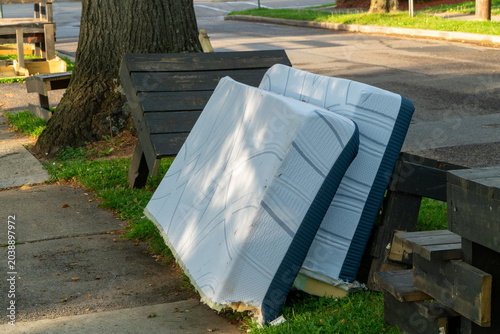 Tableau sur toile Two discarded mattresses leaning against a wooden bench outdoors on a grassy verge next to a sidewalk and tree, symbolizing urban waste, environmental pollution, and the challenge of bulk trash