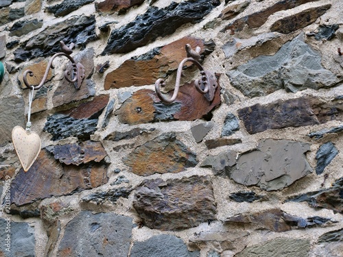 A close-up shot of a rustic medieval stone wall featuring two ornate, rusty
wrought iron hooks shaped like birds. Hanging from the left hook is a small,
fabric heart decoration with a dotted pattern. 