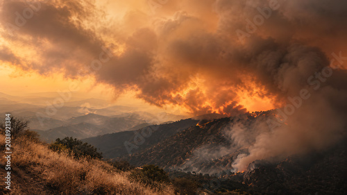 Aerial view of a massive wildfire burning through a mountain forest, creating thick smoke and glowing orange flames at sunset