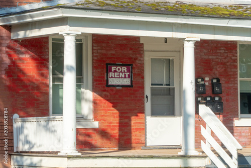 Tableau sur toile Historic red brick apartment building with a prominent 'FOR RENT' sign on its inviting shaded porch, featuring classic white columns, multiple mailboxes, and a moss-covered roof, perfect for real