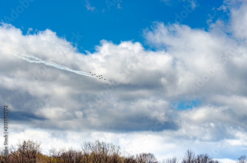 Nine fighter jets in formation flying through clouds above a treeline