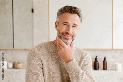 Healthy looking older man in his fifties with neatly styled beard, smiling confidently in modern bathroom setting. warm tones and natural materials create welcoming