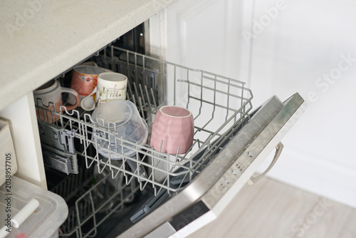 Open Dishwasher Lower Rack Loaded With Cups Mugs Bowls And Plastic Containers Inside Kitchen Under Countertop Showing Metal Basket And Assorted Tableware.