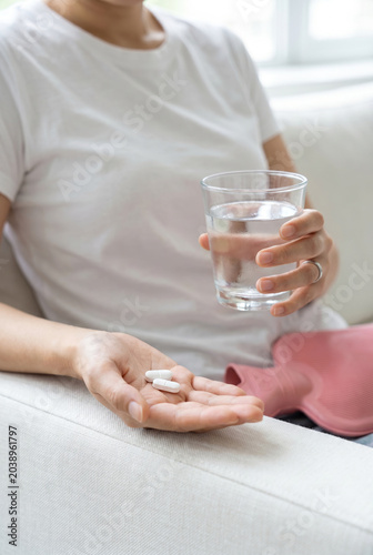 Close up of woman hands holding pills and glass of water with hot water bottle bag on sofa, period cramps or stomach ache concept.