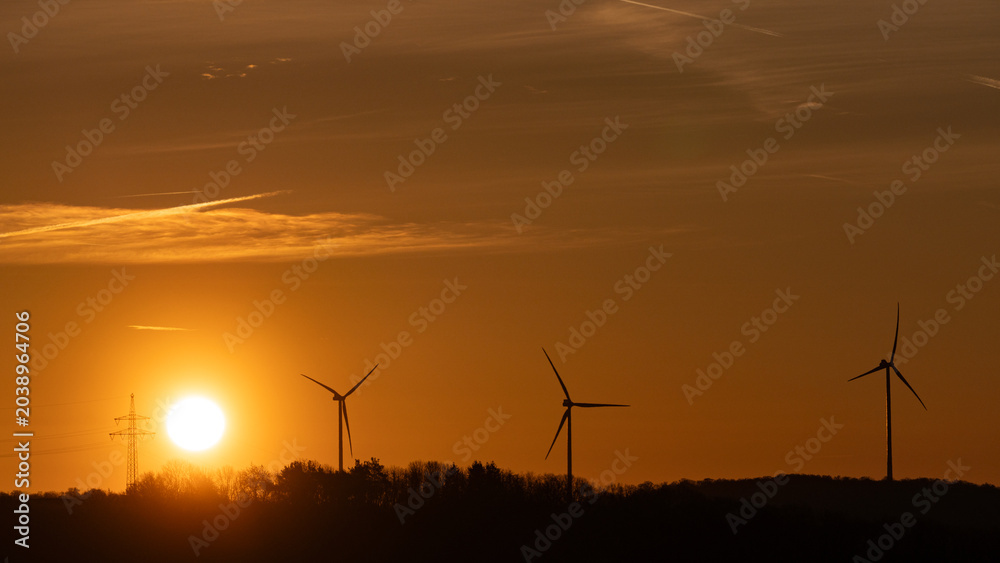 Obraz premium Wind turbine silhouettes against dramatic orange golden sunset sky with glowing sun and electricity pylon, renewable energy concept