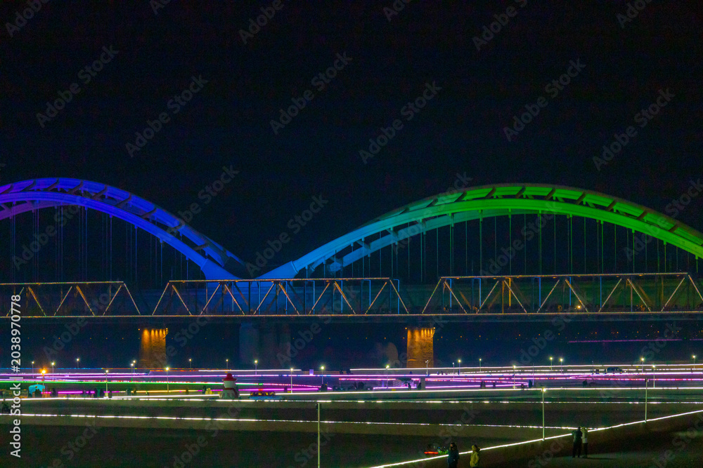 Fototapeta premium Night view of an illuminated city bridge over river with skyline and water reflections in Harbin, China.