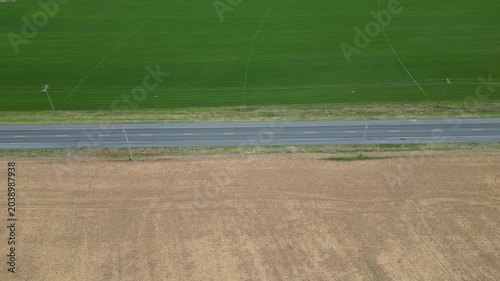 Low-altitude drone perspective of agricultural fields separated by a rural road, featuring vibrant green farmland on one side and light yellow cultivated soil prepared for planting on the other