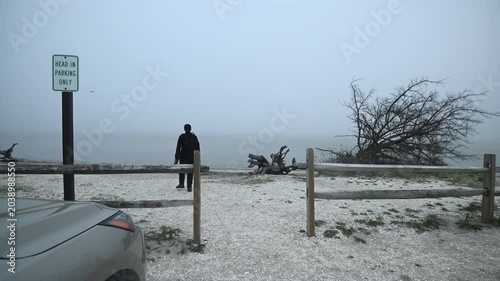 A person dressed in black and carrying a camera backpack walks through a beach access fence toward the shoreline at Assateague Island, Maryland. Dense sea fog blankets the coast, creating a mysterious