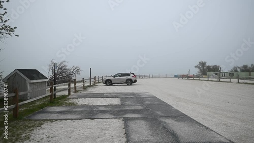 Side-view cinematic footage of an SUV slowly pulling into a fenced parking area at Assateague Island, Maryland. Dense fog blankets the nearby beach as a person dressed in black steps out of the vehicl