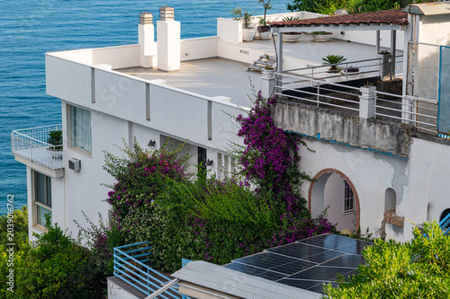 High-angle view of a white luxury villa with a spacious rooftop terrace, solar panels, and blooming purple bougainvillea overlooking the blue Mediterranean sea.