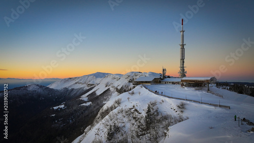 Aerial view of Col de la Faucille featuring the telecommunications tower and snow-covered mountain ridges under a sunset sky in Gex, Auvergne-Rhone-Alpes, France.