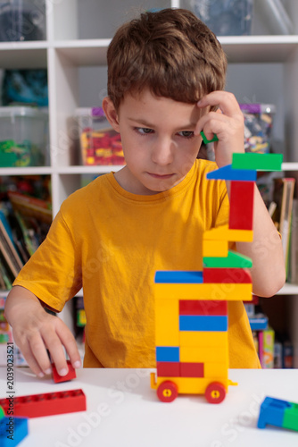 Thinking boy pausing during block tower construction, thinking child with hand near head deciding next step, thinking preschooler solving building challenge in kindergarten room