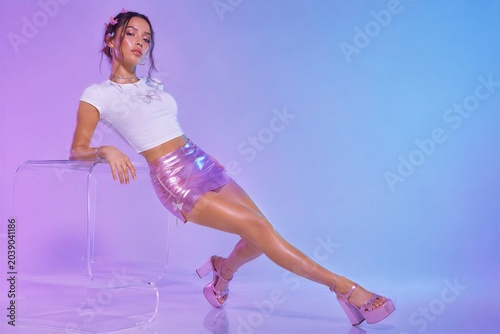 Young woman posing in stylish Y2K fashion aesthetic. Girl wearing metallic purple miniskirt and butterfly white crop top in studio. Retro 2000s trend, nostalgic pop culture clothing look