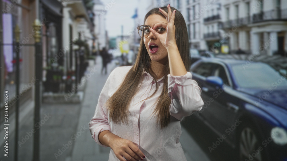 Fototapeta premium Woman wearing glasses making circle gesture over eye while touching chest, posed on a street beside a parked blue car and storefronts; playful curiosity.