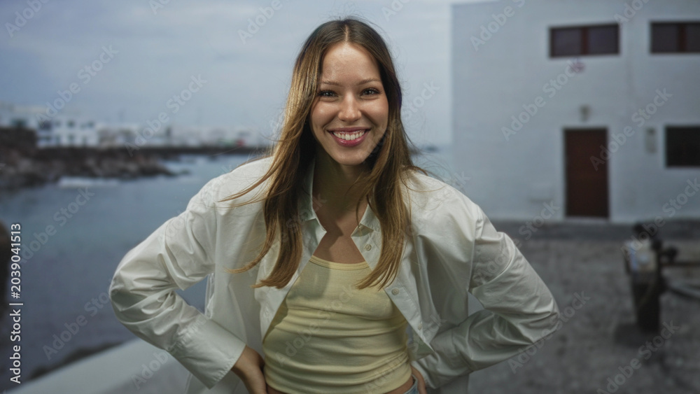 Fototapeta premium Woman smiling with hand on hip on street beside seaside promenade, leaning forward in a casual tank top; carefree joy.