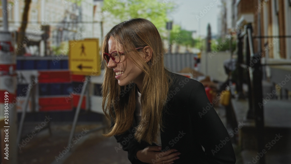 Fototapeta premium Woman clutching stomach with hand and wincing on street near barricade; distress pain urgency emergency.