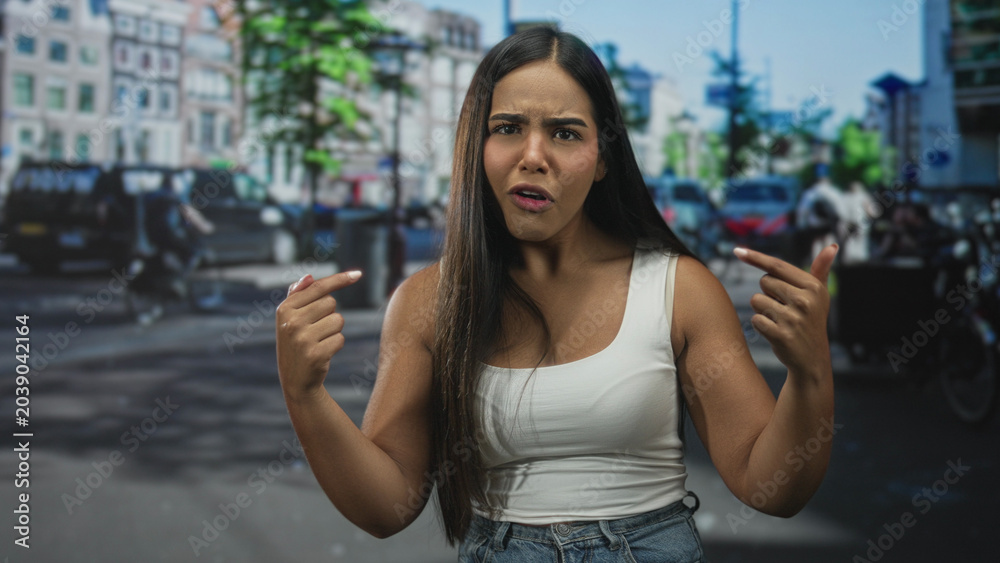Fototapeta premium Hispanic woman points fingers at her chest with bare arms visible on a busy street; confusion doubt.