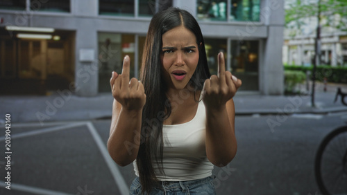 Foto Hispanic woman raises both middle fingers toward camera with scowl while standing on city street in front of office building wearing white tank top and jeans; defiance