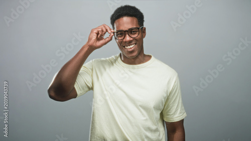 Foto Black man adjusts eyeglasses with right hand while smiling, wearing pale t shirt against studio grey wall; confidence friendliness