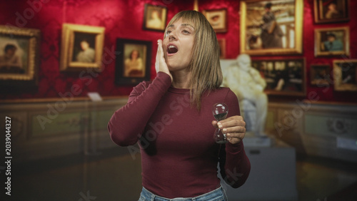 Tableau sur toile Woman holding hourglass and touching cheek in museum building surrounded by framed paintings and statue; time passing reflection