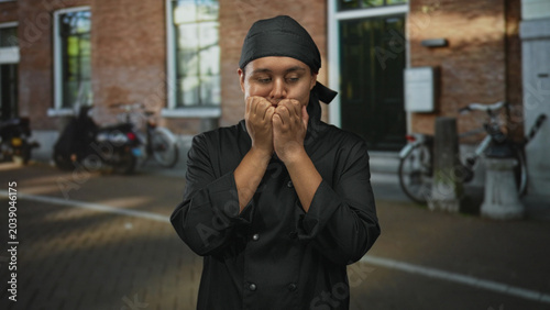 Canvas-taulu Man chef in black uniform with hand to chin on street near brick building and parked bicycles; quiet thinking