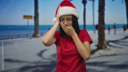 Cuadro en lienzo Woman clutching head with both hands on seaside street promenade, wearing santa hat and red shirt; stress