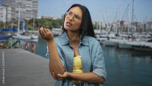 Foto Woman wearing glasses points finger and holds forearm while standing on a marina pier near moored boats; disapproval assertive communication