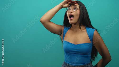 Foto Woman with glasses shielding her eyes with one hand, mouth open in a teal studio while looking into the distance; surprise