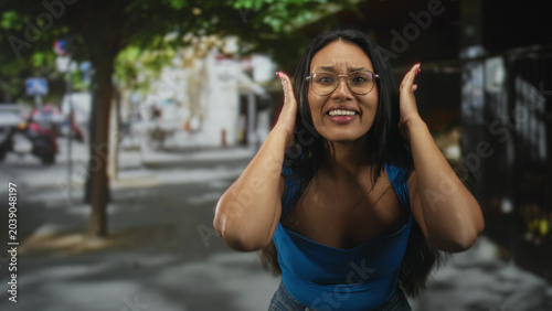 Canvastavla Woman with hands at her head and mouth open showing tongue, wearing blue top and glasses, reaching toward camera on a street; exasperation moment