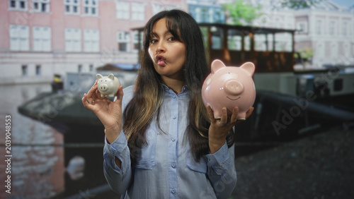 Konstfotografi Brunette hispanic woman holding a large pink piggybank and a small white piggy with both hands on an amsterdam street near a canal; playful savings comparison