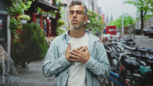 Foto Man with glasses clutching chest with both hands on a city street beside parked bicycles and a cafe storefront; concern heartattack pain
