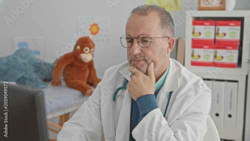Canvas Print Man doctor, middle age, wearing glasses and stethoscope, hand on chin at computer in pediatric clinic with stuffed monkey on exam couch; thoughtful concern