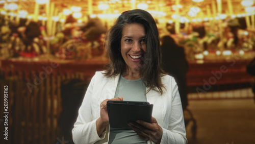 Konstfotografi Brunette middle age woman holding a tablet, tapping and scrolling the screen while smiling then frowning amid bright carousel lights in a building; curiosity learning